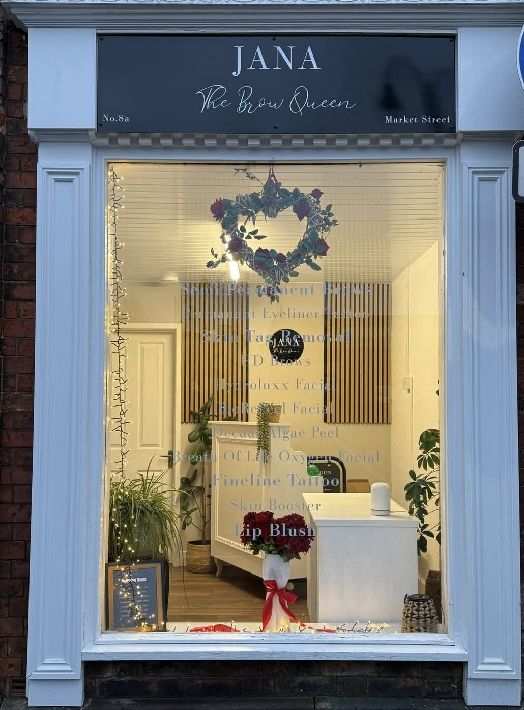The shop at night, its window lit and the JANA The Brow Queen sign above